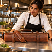 A woman in a white chef coat and black apron smiles while leaning over a stainless steel kitchen counter next to a large brown leather travel case with a shoulder strap.
