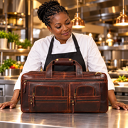 A woman in a white chef's coat and black apron stands behind a large brown leather travel bag with gold-tone hardware in a professional kitchen setting.